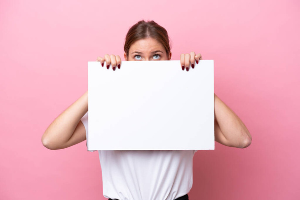Young caucasian woman isolated on pink background holding an empty placard and hiding behind it - Photo, image