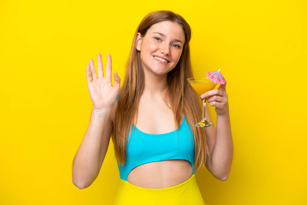 Joven mujer caucásica sosteniendo un cóctel aislado sobre fondo amarillo saludando con la mano con expresión feliz - Foto, Imagen