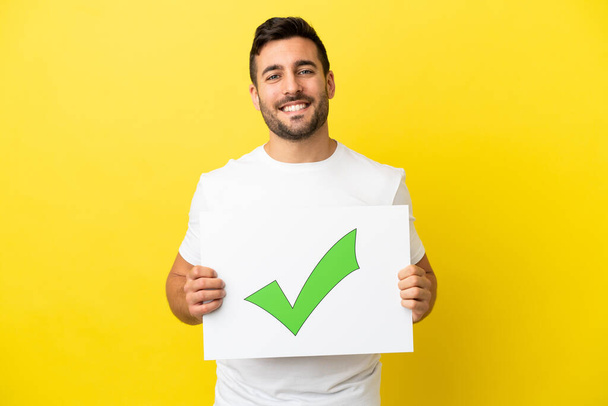 Young handsome caucasian man isolated on yellow background holding a placard with text Green check mark icon with happy expression - Photo, Image