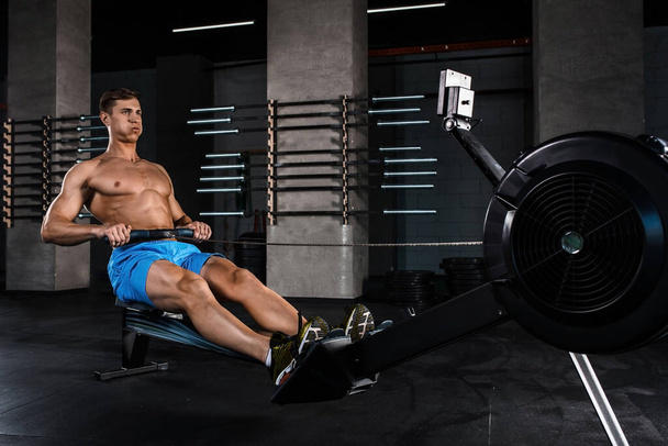 Culturista trabajando en sus piernas con una máquina de pesas en el gimnasio. Musculoso joven en pantalones cortos azules haciendo ejercicios. - Foto, Imagen