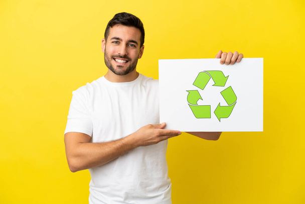 Junger gutaussehender kaukasischer Mann auf gelbem Hintergrund, der ein Plakat mit einer Recycling-Ikone mit glücklichem Gesichtsausdruck hält - Foto, Bild