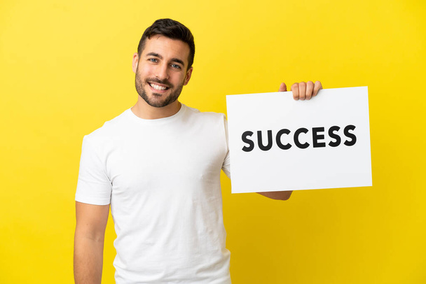 Young handsome caucasian man isolated on yellow background holding a placard with text SUCCESS with happy expression - Photo, Image