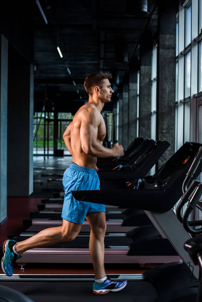 Side view full length of young man in sportswear running on treadmill at gym. Muscular young man in blue shorts doing exercises. motion blur - Photo, Image