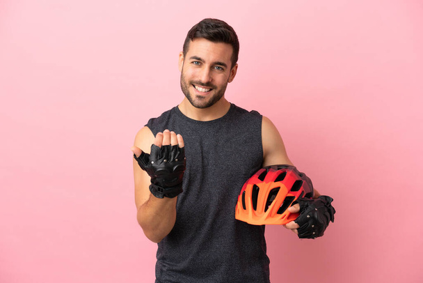 Young cyclist man isolated on pink background inviting to come with hand. Happy that you came - Foto, afbeelding