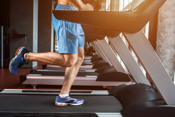 Legs running on the treadmill close up. A muscular man in yellow sneakers and blue shorts running down the treadmill in the gym. Motion blur - Zdjęcie, obraz