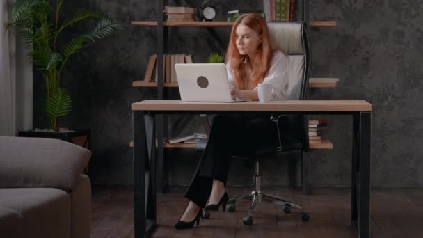 červená hlava žena psaní na notebook pití kávy sedí na stole doma moderní domácí kancelář v bytě - Záběry, video
