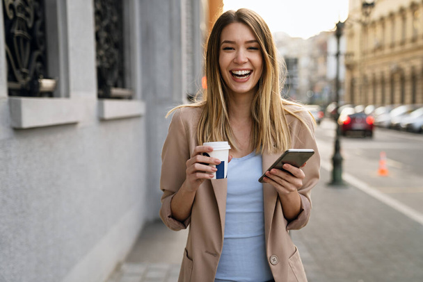 Beautiful young success woman with coffee using mobile phone against urban city background. People business break device concept - Foto, Imagen