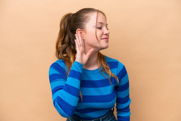 Young caucasian woman isolated on beige background listening to something by putting hand on the ear - Foto, afbeelding