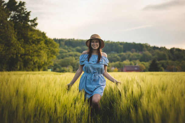Lélegzetelállító, őszinte portré egy 20-24 éves barnáról, aki gyönyörű kék ruhában és kalapban sétál a kukoricaföldön, természetesen mosolyogva. Divat vintage stílus. Egy barna hajú európai nő természetes szépsége. - Fotó, kép