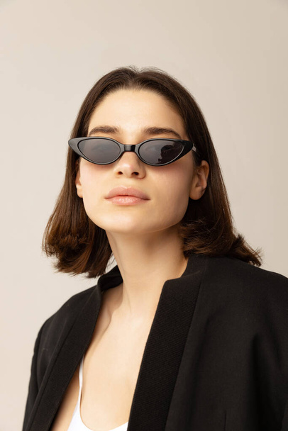 One young woman in black jacket, white top and trendy sunglasses posing, looking at camera isolated over grey background. Expressing style. Concept of style, retro, fashion, youth, beauty, emotions - Foto, immagini