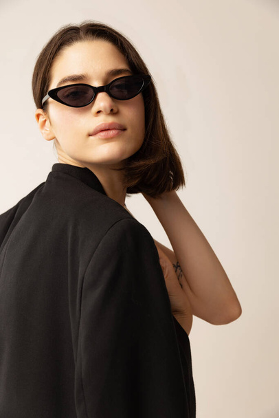 Retrato de mujer joven con estilo en chaqueta negra y gafas de sol de moda posando aisladas sobre fondo gris estudio. Feminidad. Concepto de estilo, retro, moda, juventud, belleza, emociones - Foto, Imagen