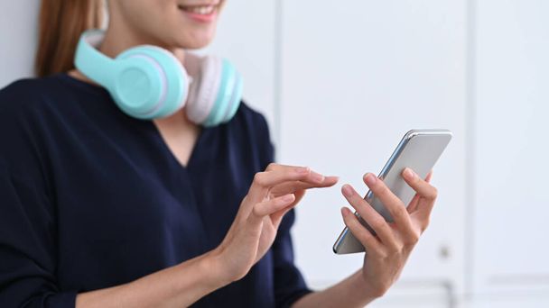Smiling young woman with wireless headphones using smart phone in modern home. - Photo, Image