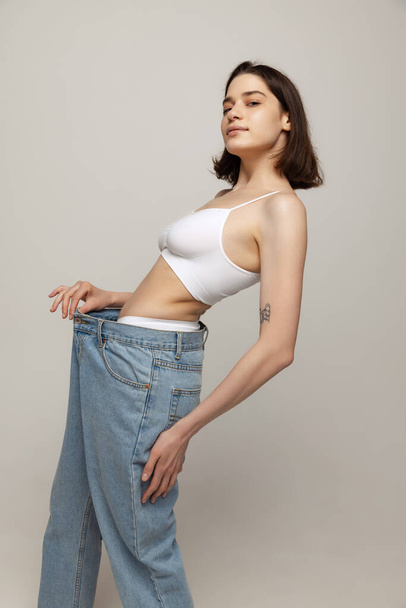 Portrait of young slim wom,an posing in white top and oversized jeans isolated oevr grey studio background. Weight-loss. Concept of beauty, body and skin care, health, spa, cosmetics, ad - Foto, Bild