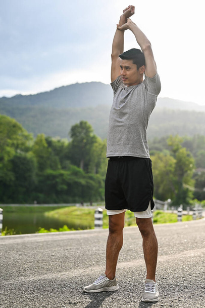 Портрет здорового азіатського чоловіка в спортивному одязі, розтягує руки, розігрівається, готується до бігу в прекрасному парку зелені. Спорт активує концепцію
. - Фото, зображення