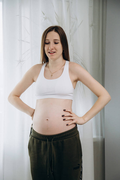 妊娠中の長髪の女の子で緑のパンツは窓の近くに立つ - 写真・画像