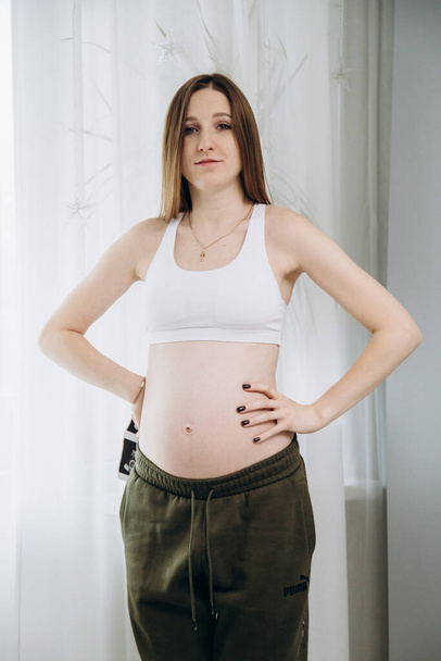Terhes hosszú hajú lány zöld nadrágban áll az ablak mellett - Fotó, kép