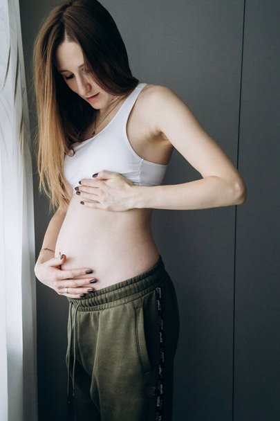妊娠中の長髪の女の子で緑のパンツは窓の近くに立つ - 写真・画像