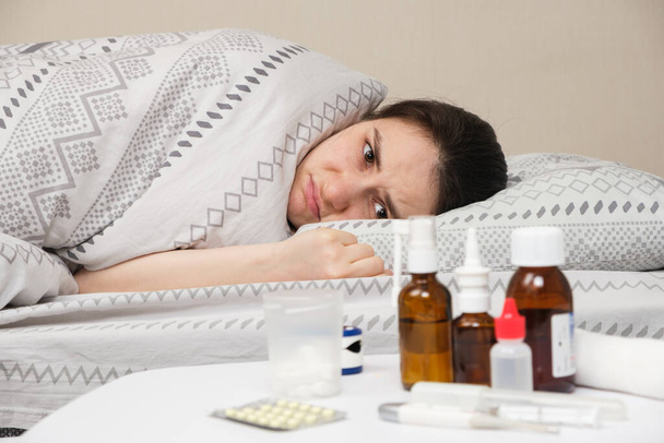 A sick woman lies in bed and looks sadly at the bedside table with medicines. - Фото, зображення