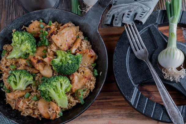 Healthy fitness meal with pan fried and chopped chicken breast, brown rice and broccoli. Served in a rustic iron pan on wooden table background with dumbbell. Top view - Photo, Image