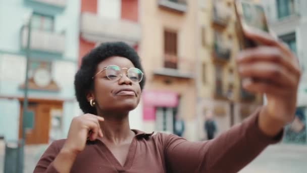 Retrato de la alegre mujer rizada afroamericana tomando selfie en la calle. Guapa blogger mostrando gesto de paz tomando fotos al aire libre - Metraje, vídeo