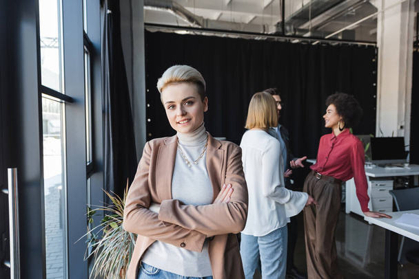 happy businesswoman standing with crossed arms while multiethnic colleagues talking in advertising agency - Fotografie, Obrázek