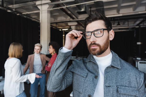 бородатий бізнесмен коригує окуляри і дивиться в сторону, поки багатокультурна команда розмовляє на розмитому фоні
 - Фото, зображення