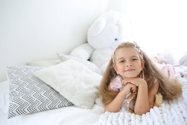 Little cute Caucasian girl with a smile lying on the bed. Bright interior in the children's room. Modern nursery. Copy space on the left - Foto, Imagem