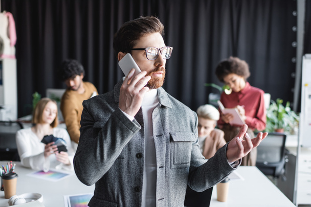 advertising manager in eyeglasses talking on smartphone near blurred multicultural colleagues  - Фото, изображение