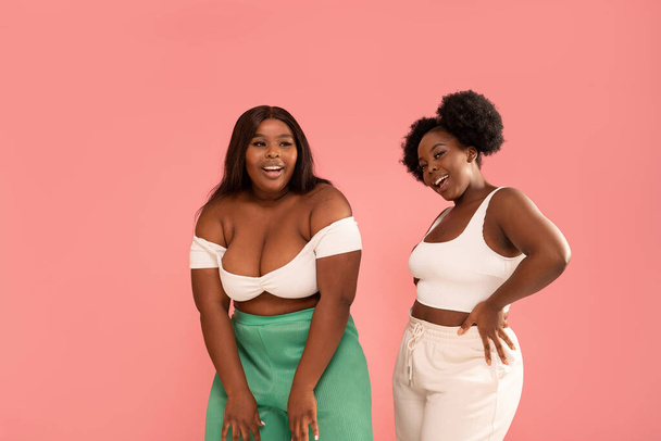 Studio shot of happy dark skinned, young sisters enjoying good moment, dancing and having fun together, posing over pink pastel studio background. Friendship and real people emotions concept. Body positive. Copy space. - Foto, imagen