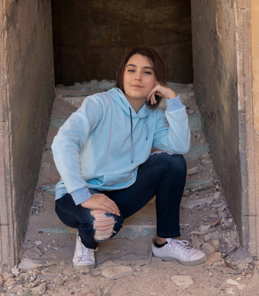 Tiener meisje in lichtblauwe hoodie zittend op haar haunches vlakbij een muur. Tiener meisje brengt tijd alleen door buiten. Kleding model. Adolescentieconcept - Foto, afbeelding