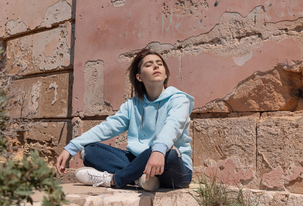 Teenager girl in light blue hoodie sitting on her haunches with eyes closed near a wall. Serious Teen girl spending time outdoor alone. Clothing mockup. Adolescence concept - Valokuva, kuva
