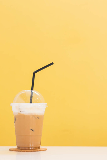 Geeister Cappuccino mit Eis, das mit weißem Milchschaum belegt und mit einem Plastikdeckel bedeckt ist, der auf einem Tisch im Café auf einem leuchtend gelben, verschwommenen Hintergrund steht. Für das Einfügen von Text steht Kopierraum zur Verfügung - Foto, Bild