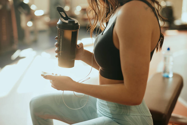 Cropped picture of a strong sportswoman sits in a gym, drinks protein drink and using a phone while drinking protein drink and taking a break. - Fotografie, Obrázek