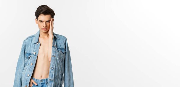 Handsome and sassy gay man wearing denim jacket on bare torso, touching his face and looking confident at camera, standing over white background. - Фото, изображение