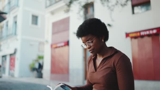 Gözlüklü güzel Afrikalı Amerikalı kadın sokakta oturmuş kitap okuyor. Açık havada okuyan esmer tenli bir kız. - Video, Çekim