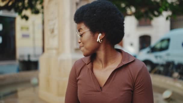 Csinos afrikai lány szemüveges élvezze a jó zenét vezeték nélküli fülhallgató a szabadban. Vonzó sötét bőrű nő vár barát az utcán - Felvétel, videó