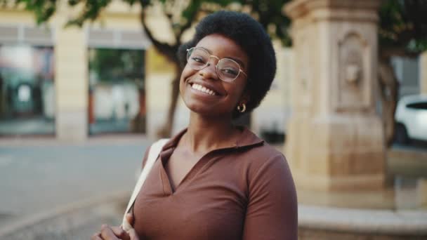 Schöne Afrikanerin mit Brille, die charmant auf der Straße in die Kamera posiert. Unbekümmert lockiges Mädchen, das glücklich im Freien aussieht - Filmmaterial, Video