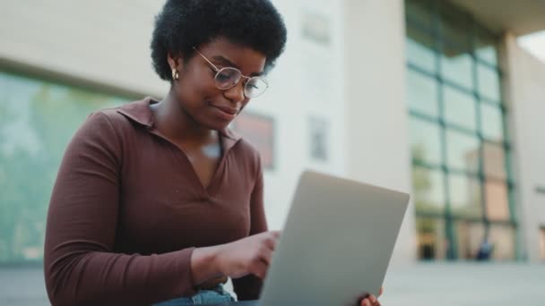 Giovane imprenditrice digitando e-mail di lavoro sul computer portatile all'aperto. Studente afroamericano ragazza in occhiali lettura lezione utilizzando il computer portatile sulla strada - Filmati, video