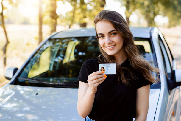 Aantrekkelijke jonge vrouw in zwart T-shirt voor de moderne auto opscheppen over het ontvangen van een rijbewijs met rijbewijs aan de camera en zich gelukkig voelen - Foto, afbeelding