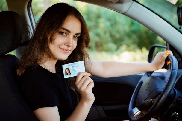 Junges Mädchen mit schönem Lächeln sitzt im Auto und zeigt der Kamera ihren Führerschein. Frau hat Führerschein und fühlt sich glücklich und aufgeregt - Foto, Bild