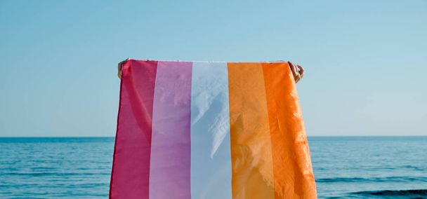 closeup of a woman, seen from behind, holding a lesbian pride flag on the air above her head facing the sea in a sunny day - Fotó, kép