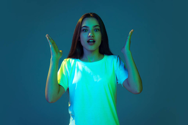 Beautiful surprised girl with long loose hair looking at camera with open mouth isolated on blue background in neon light. Concept of beauty, art, fashion, youth, sales and ads. Student looks excited - Foto, afbeelding