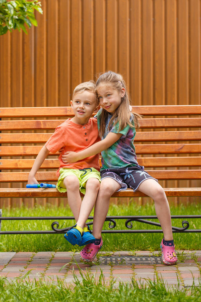 Мальчик и девочка сидят на скамейке. В солнечный летний день пятилетний мальчик и девочка сидят на скамейке в саду. - Фото, изображение