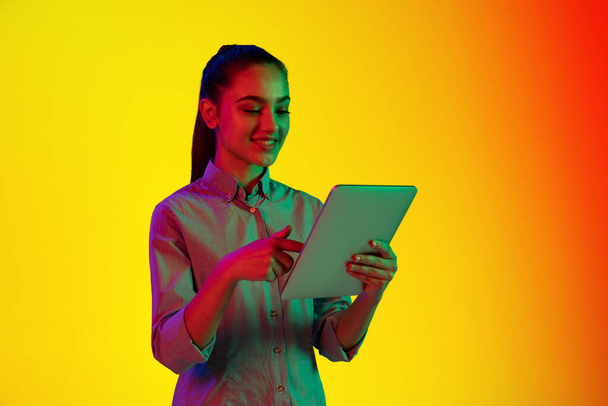 Portrait of young girl using tablet isolated over gradient yellow red background in neon light. Concept of emotions, facial expression, youth, aspiration. Copy space for ad - Foto, afbeelding