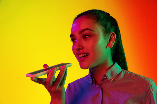 Closeup portrait of young smiling girl using phone isolated over gradient yellow red background in neon light. Concept of emotions, facial expression, youth, aspiration. Copy space for ad - Foto, afbeelding