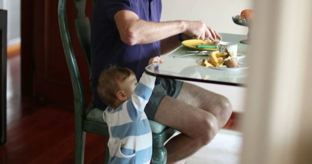Candid baby standing next to grandpa while eating morning breakfast - Fotó, kép