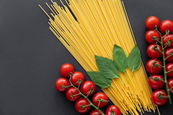 Top view of raw spaghetti near cheery tomatoes and basil on black background  - Zdjęcie, obraz