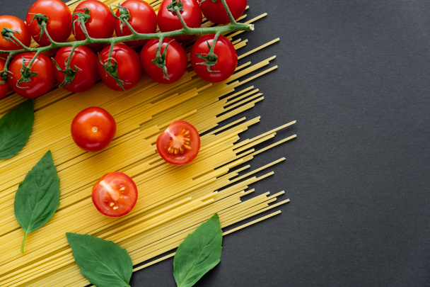 Top view of organic tomatoes and basil on raw spaghetti on black background  - Foto, afbeelding