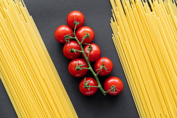 Widok z góry pomidory wiśniowe na gałęzi między surowe spaghetti na czarnym tle  - Zdjęcie, obraz