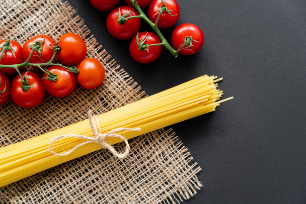 Top view of raw spaghetti and cherry tomatoes on sackcloth on black background  - Photo, Image